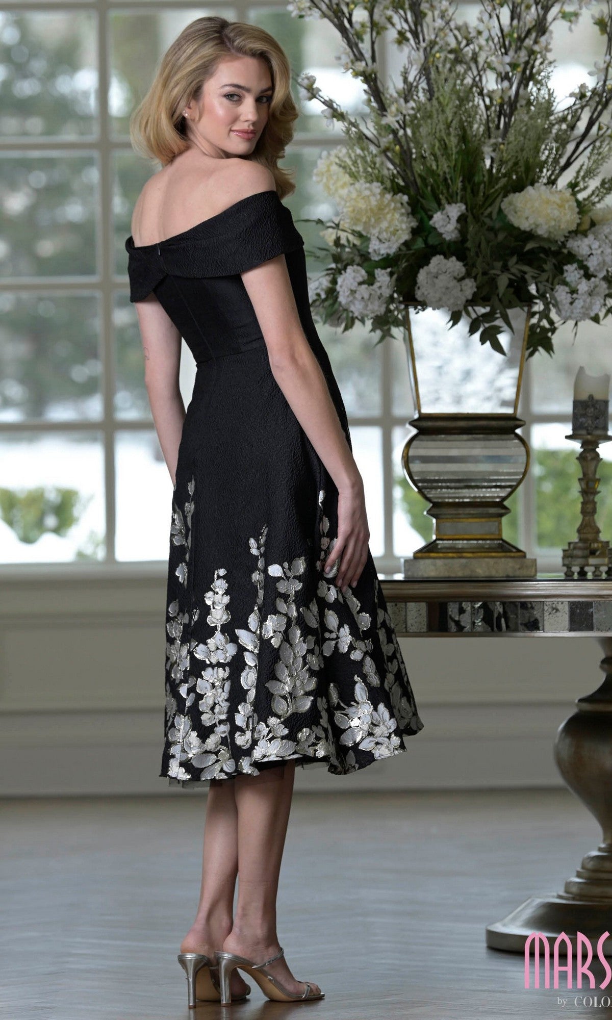 A woman stands indoors near a table with large floral arrangements, wearing the Marsoni MV1414—a stunning knee-length semi-formal black dress with off-the-shoulder silver floral patterns.
